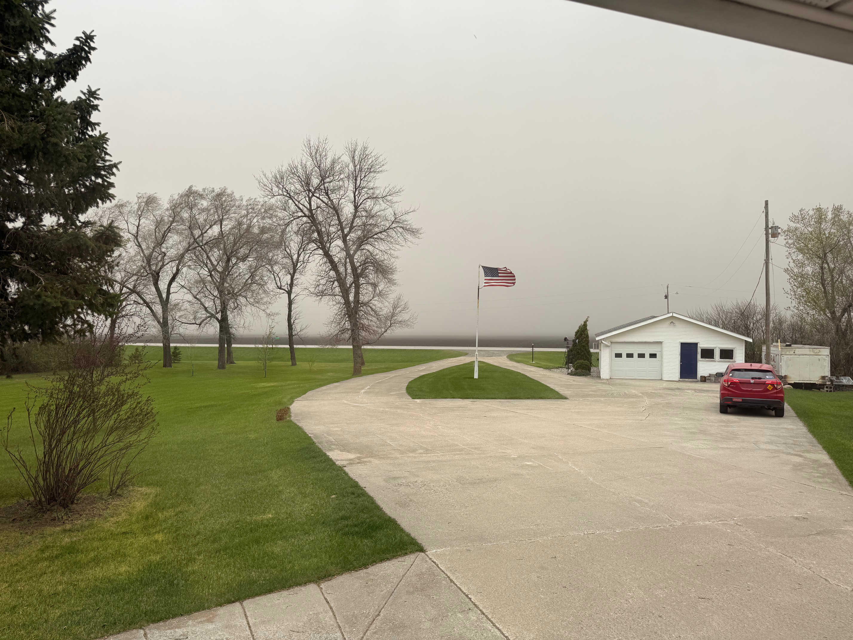 A driveway leads to a small white building and a parked red car, with an American flag flying nearby and bare trees in the background under a dusty sky.